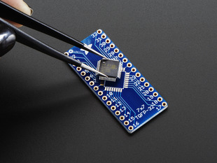 A pair of tweezers holding a microchip hovers over the SMT Breakout PCB for 32-QFN or 32-TQFP placement on a long blue rectangular breakout board.