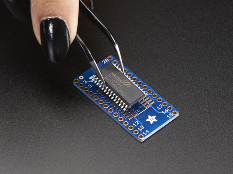 A pair of tweezers holding a microchip hovers over the SMT Breakout PCB for SOIC-28 or TSSOP-28.