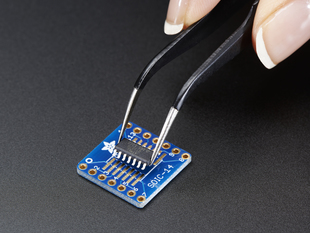 A pair of tweezers holding a microchip hovers over the SMT Breakout PCB for SOIC-14 or TSSOP-14.