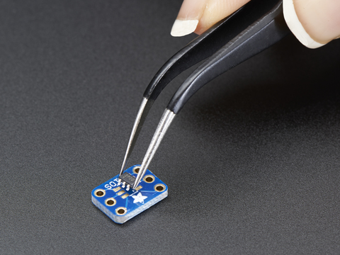 A pair of tweezers holding a microchip hovers over a small rectangular Breakout PCB.