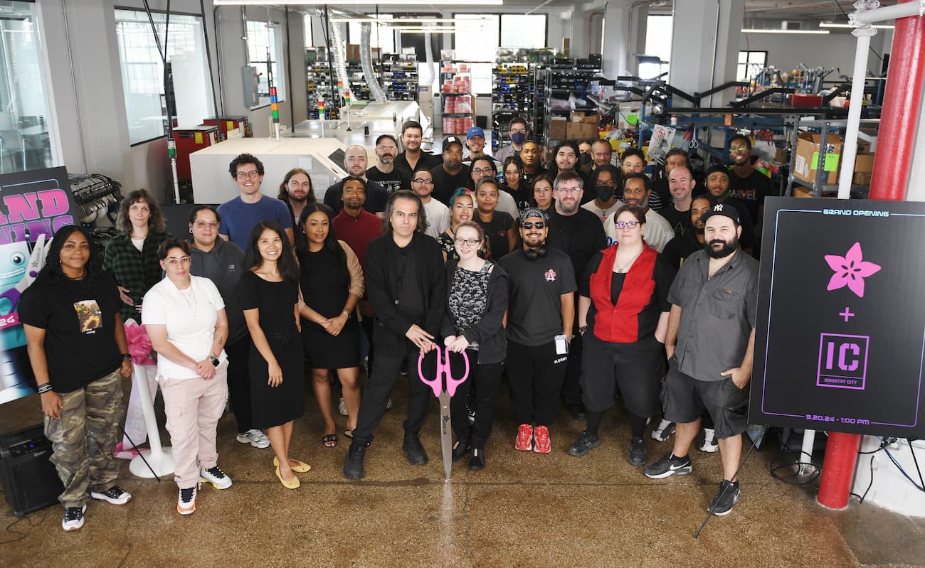 Group photo of approximately 40 people in front of manufacturing equipment. There is a pair of giant pink scissors.